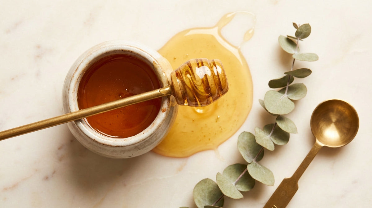 Overhead flat-lay of golden manuka honey dipper on cream marble surface with measuring spoon